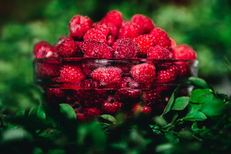 Foto framboesas em uma tigela de vidro com galhos e folhas em um escuro. fruta saudável e sazonal para comer.