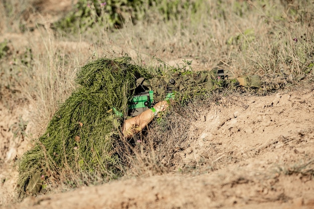 Francotirador armado con rifle de francotirador de gran calibre, disparando objetivos enemigos a distancia del refugio, sentado en una emboscada.