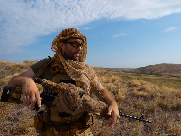 Francotirador mercenario con ropa de camuflaje bajo el sol abrasador se encuentra con un rifle y inspecciona