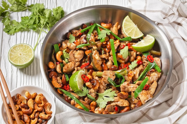 Frango de caju tailandês salteado em uma panela sobre uma mesa de madeira com pauzinhos e ingredientes vista horizontal de cima