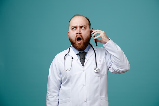 Franzindo a testa jovem médico masculino vestindo jaleco e estetoscópio em volta do pescoço, olhando para o lado enquanto fala no telefone isolado no fundo azul