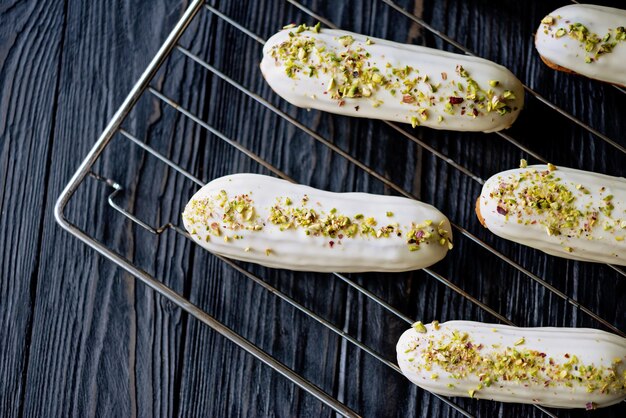 Französische Dessert-Eclairs oder Profiteroles mit weißer Schokoladenglasur mit Pistazien, auf einem Gebäckgestell. Gebäckkuchen mit Vanillepudding, Pistaziencreme und Belag. Weicher selektiver Fokus.