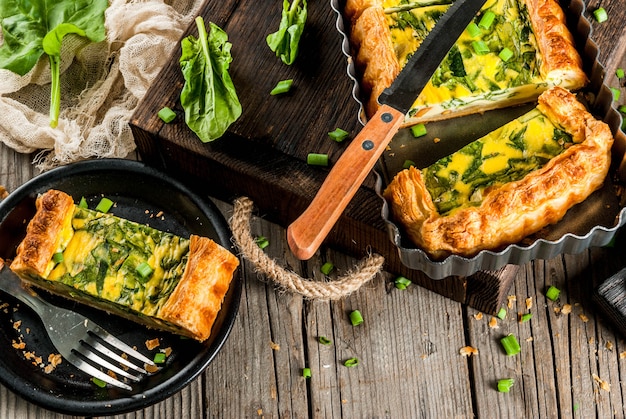 Französische Hausmannskost. Kasserolle. Kuchen. Quiche Lorraine aus Blätterteig, mit jungen Frühlingszwiebeln und Spinat. Auf alter hölzerner rustikaler Tabelle. Schnitt. In Form zum Backen. Teller, Gabel, Messer. Copyspace Draufsicht