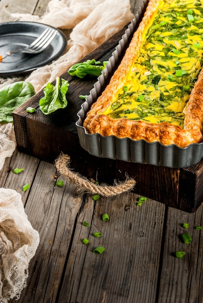 Französische Hausmannskost. Kasserolle. Kuchen. Quiche Lorraine aus Blätterteig, mit jungen Frühlingszwiebeln und Spinat. Auf alter hölzerner rustikaler Tabelle. Schnitt. In Form zum Backen. Teller, Gabel, Messer.