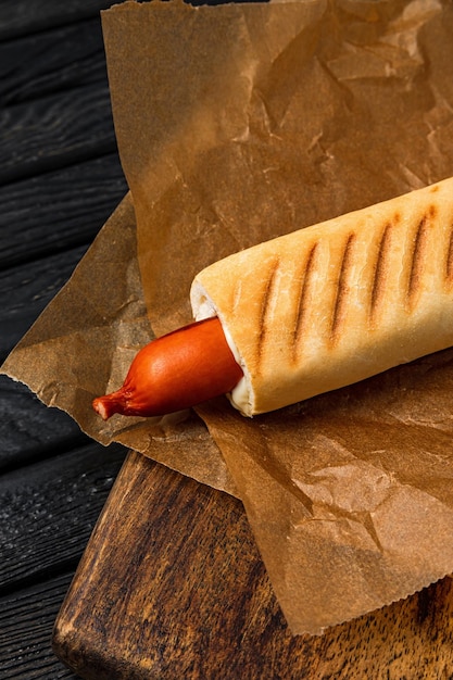 Französischer Hotdog in einem knusprigen Brötchen, serviert auf Pergamentpapier auf Holzbrett auf dunklem Holzhintergrund, Nahaufnahme