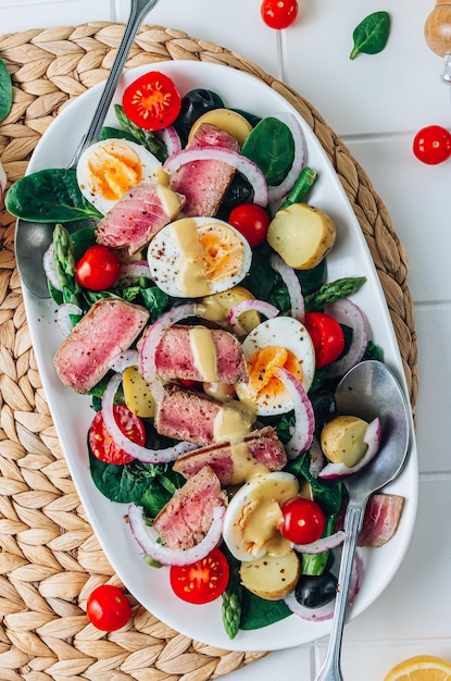 Französischer Salat Nicoise mit Thunfischsteak, Spargel, Tomaten, Eiern, Kartoffeln und schwarzen Oliven auf einem weißen Ba