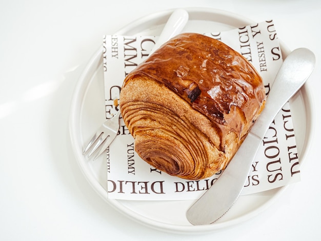 Französisches Croissant mit Metallmesser und -gabel auf Papier auf weißer runder Keramikplatte isoliert auf weißem Tischhintergrund mit natürlichem Sonnenlicht Frisch gebackenes Schokoladencroissant-Rezept
