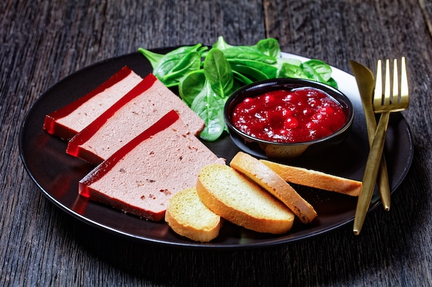 Französisches Gericht Entenleberpastete mit Preiselbeermarmelade auf einem schwarzen Teller mit Preiselbeersauce, Spinat und Brioche-Croutons, mit goldenem Besteck auf einem dunklen Holztisch, Nahaufnahme, Draufsicht