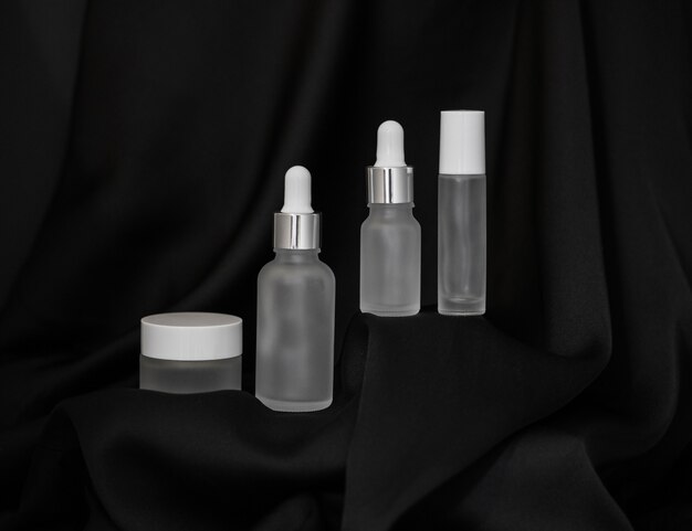 Frasco cosmético, spray e dois frascos de cosméticos com um conta-gotas em um fundo de seda preta, spray cosmético e um porta-frasco pequeno na plataforma acima