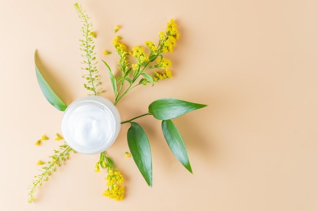 Frasco de creme aberto para cuidados com a pele com folhas verdes e flores em fundo bege closeup foto minimalista para apresentação de produtos de primavera