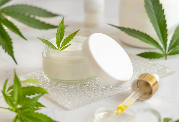 Frasco de creme branco aberto com tampa perto de folhas de cannabis Maquete cosmética