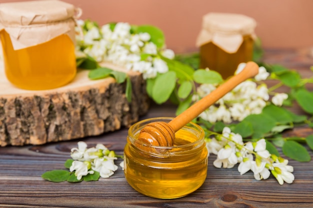 Frasco de mel doce cercado de flores de acácia da primavera O mel flui de uma colher em uma jarra de mel de acácia fresco claro em fundo de madeira