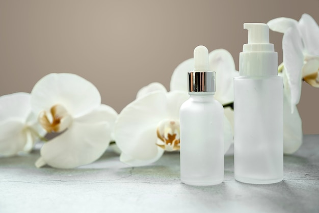 Frasco de soro de vidro branco e frasco de bomba de loção com maquete de dispensador no banheiro com flores de orquídea ao fundo, produtos cosméticos sem marca, maquete de marca de produto cosmético de spa