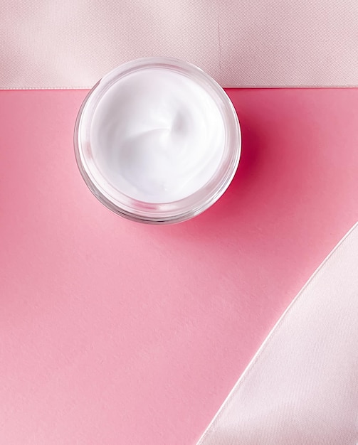 Frasco hidratante de creme facial de cosméticos para a pele e fita de seda branca em fundo rosa produto de beleza plano fechado