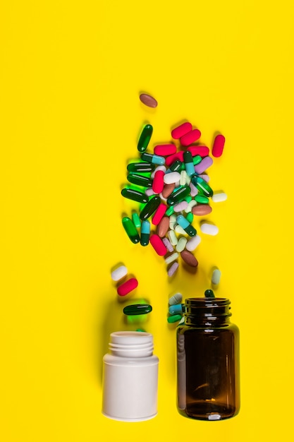 Un frasco marrón vacío con una tapa blanca cerrada y un frasco blanco de medicamentos se encuentran junto a una gran cantidad de tabletas y cápsulas.