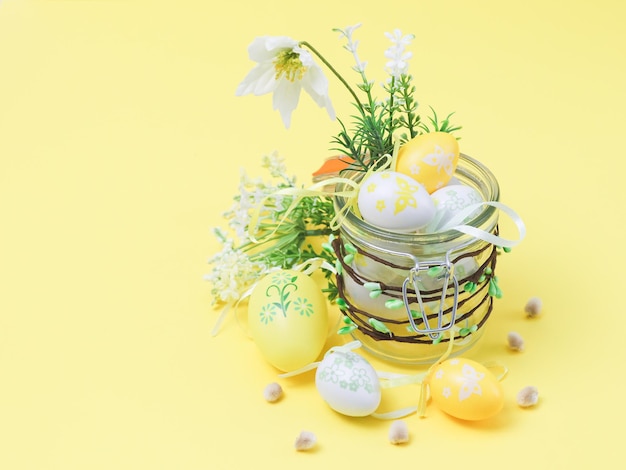 Un frasco de vidrio con brotes en los que hay huevos de Pascua decorativos, flores de primavera y brotes de sauce en un amarillo