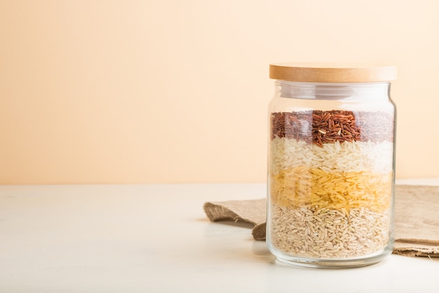 Frasco de vidrio con diferentes tipos de arroz vertido en capas sobre fondo blanco y naranja. Vista lateral, copia espacio.