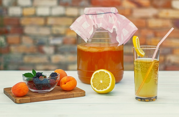 Frasco de vidrio con kombucha, un vaso vertido con kombucha con una rodaja de limón, frutas en una tabla de madera