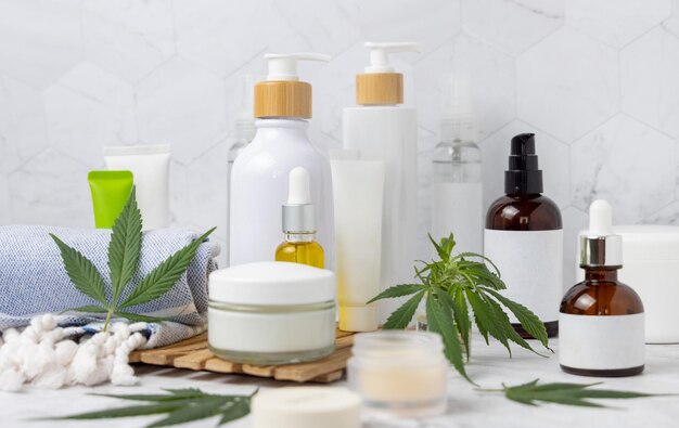 Frascos cosméticos em branco tubos e garrafas perto de folhas de cannabis verdes em bandeja de madeira na casa de banho perto de outras garrafas close up mockup produtos de beleza orgânicos de cuidados com a pele cosméticos naturais CBD
