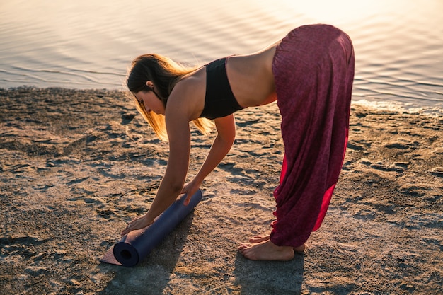 Frau am Strand bei Sonnenuntergang beginnt Yoga-Training mit Yogamatte. Morgendliches Aufwärmtraining mit natürlicher Dehnung