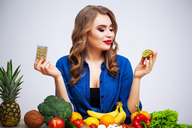 Frau an einem Tisch, der eine Kiwi und Pillen auf einem Hintergrund von Obst und Gemüse hält.