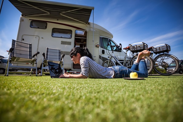 Frau auf dem Rasen, Blick auf den Laptop in der Nähe des Campingplatzes. Wohnwagen Auto Urlaub. Familienurlaubsreise, Urlaubsreise im Wohnmobil