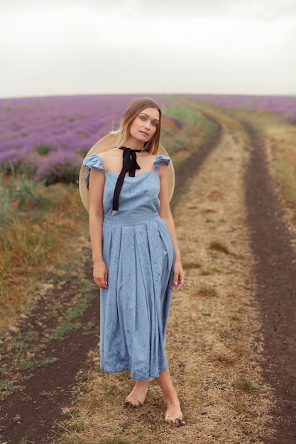 Frau auf einem schmutzigen Bodenweg in einem blauen Kleid und einem Weidenhut zwischen Lavendelfeldern in einer Provence