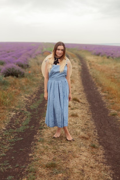 Frau auf einem schmutzigen Bodenweg in einem blauen Kleid und einem Weidenhut zwischen Lavendelfeldern in einer Provence