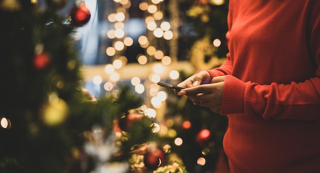 Frau auf Pullover, die auf dem Handy für Online-Messaging und soziale Kommunikation unter lebendigen Nachtlichtern einer wunderschönen Festivalparty in der Wintersaison tippt. Fügen Sie etwas Rauschen hinzu, um das Bild im Vintage-Stil anzupassen