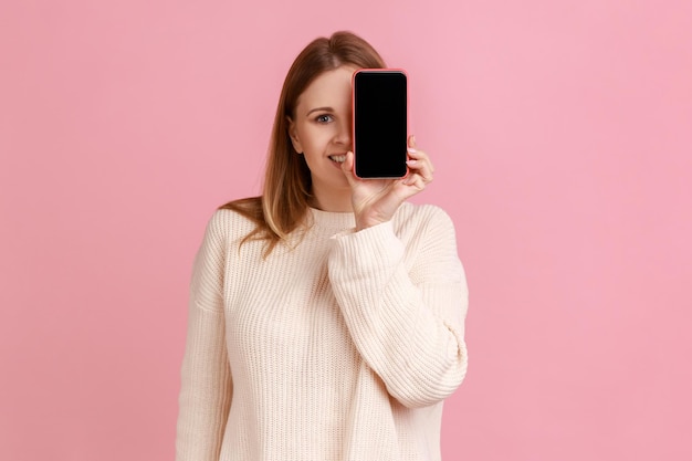 Frau bedeckt ihr Auge mit Smartphone mit leerem Display für Werbung mit spielerischem Ausdruck