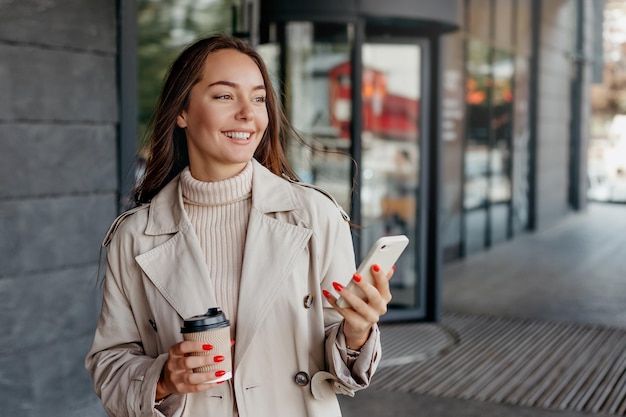 Frau benutzt ihr Smartphone, hält eine Tasse Kaffee und lächelt vor dem Hintergrund eines Bürogebäudes