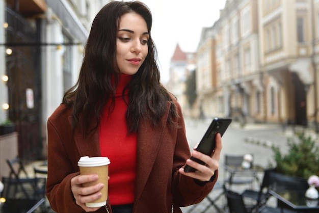 Frau benutzt Mobiltelefon draußen und hält eine Tasse Kaffee in der Hand