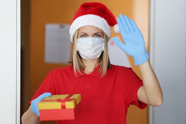 Frau Briefträger im Weihnachtsmannhut in der medizinischen Maske und in den Gummihandschuhen hält Geschenk