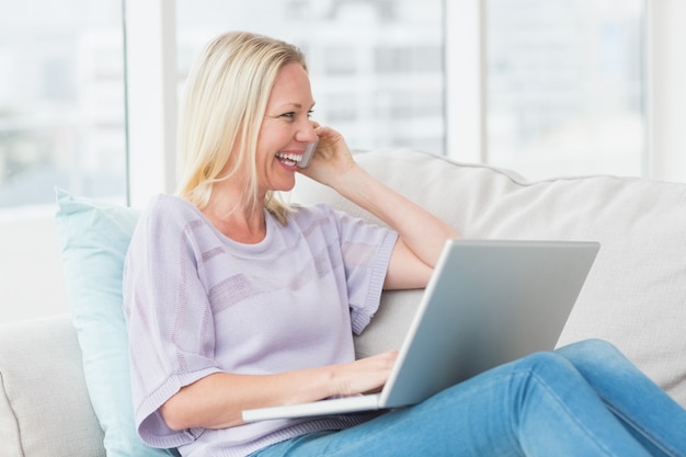 Frau, die am Handy bei der Anwendung des Laptops auf Sofa spricht