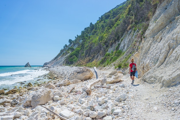 Frau, die am italienischen Strand geht. Ungleiche Küste von Conero, Kiesstrand, türkisfarbenes Wasser, echte Menschen, Rückansicht, sonniger Tag, Urlaub in Italien