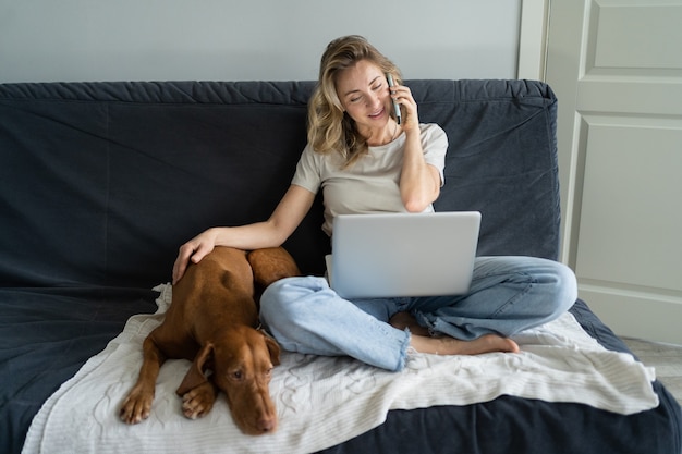 Frau, die an der Couch mit Hund sitzt, auf Handy spricht, entfernt am Computer-Laptop zu Hause arbeitet