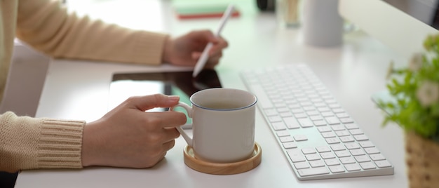 Frau, die an Tablette arbeitet und Kaffeetasse auf weißem Schreibtisch hält