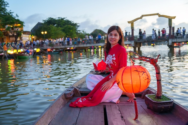 Frau, die Ao Dai vietnamesisches Kleid trägt, Sightseeing-Bootsfahrt und schwimmende Papierlaterne in der Altstadt von Hoi An, Wahrzeichen für TouristenattraktionenVietnam und Südost-Reisekonzept