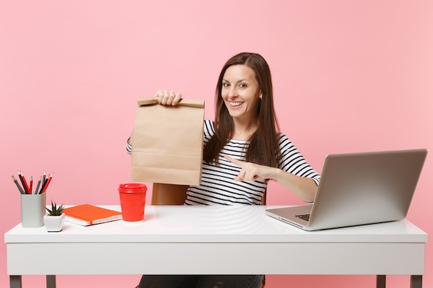 Frau, die auf braune, leere, leere Papiertüte zeigt, im Büro mit Laptop arbeiten?