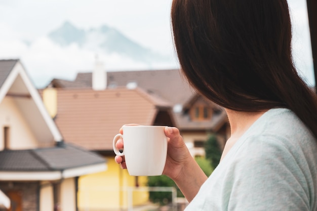 Frau, die auf dem Balkon steht und Tee oder Kaffee trinkt und erstaunlichen Blick auf die Berge genießt