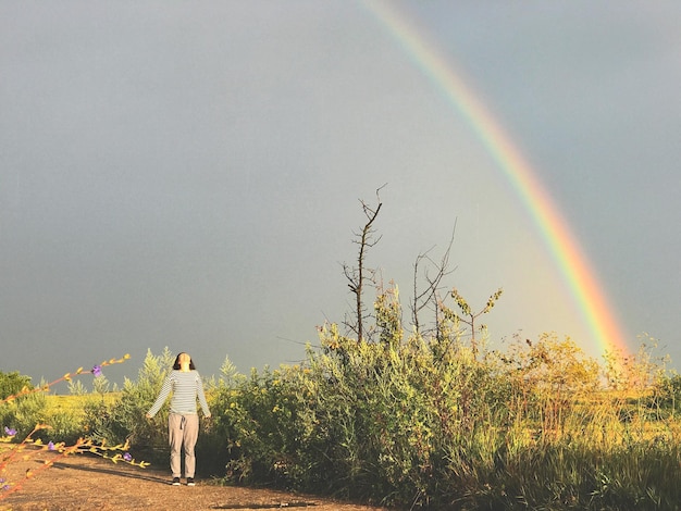 Foto frau, die auf den regenbogen schaut, während sie auf dem feld gegen den klaren himmel steht