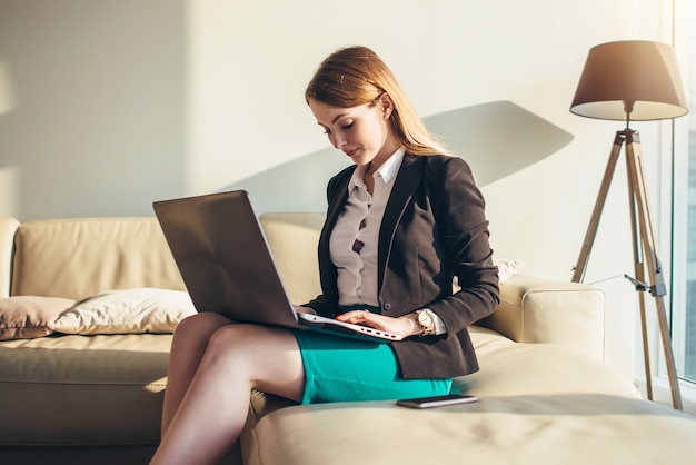 Frau, die auf einem Sofa mit einem Laptop auf ihrem Schoß sitzt, der auf Tastatur zu Hause tippt.