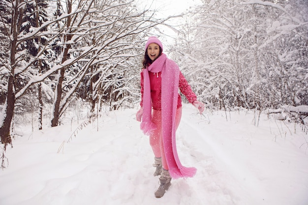 Frau, die auf einem Weg im Wald in rosa Kleidern eine Jacke einen gestrickten Schal und eine Mütze steht, steht im Winter in einem verschneiten Wald