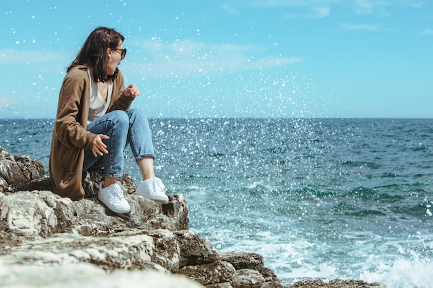 Frau, die auf einer Klippe sitzt und den Blick auf das Meer genießt, windiges Wetter, sonniger Tag