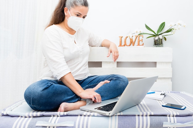 Foto frau, die auf heimsofa mit laptop mit antivirenmaske sitzt