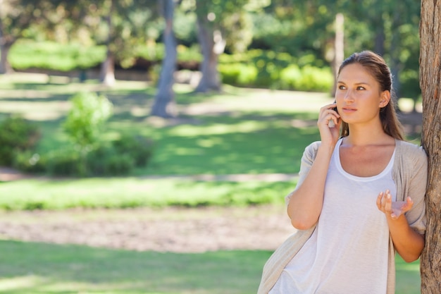 Frau, die auf ihrem Mobiltelefon beim Lehnen an einem Baum spricht