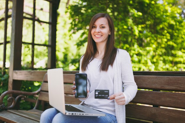 Frau, die auf Kamerakreditkarte, Handy mit leerem leerem Bildschirm zeigt, um Raum zu kopieren. Frau sitzt auf der Bank und arbeitet an einem modernen Laptop in der Straße im Freien. Mobiles Büro. Freiberufliches Geschäftskonzept.