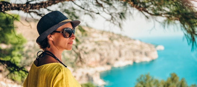 Frau, die auf Klippe unter dem Baum steht und zu einem Meer schaut