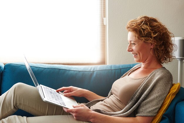 Frau, die auf Sofa mit dem Laptoplächeln liegt