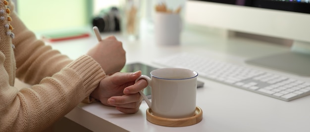Frau, die auf Tablette schaut und Kaffeetasse auf weißem Schreibtisch hält